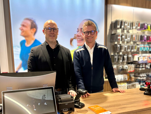 Three individuals behind a retail counter, one with curly hair and glasses, another with straight hair, and the third with short hair and glasses, with shelves of products in the background