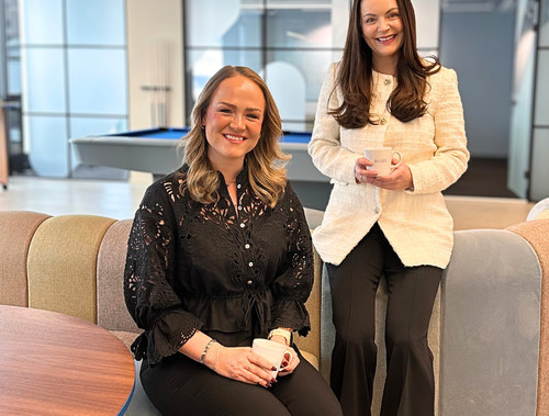 Two women sitting and standing on a sofa in a modern office setting with large windows behind them