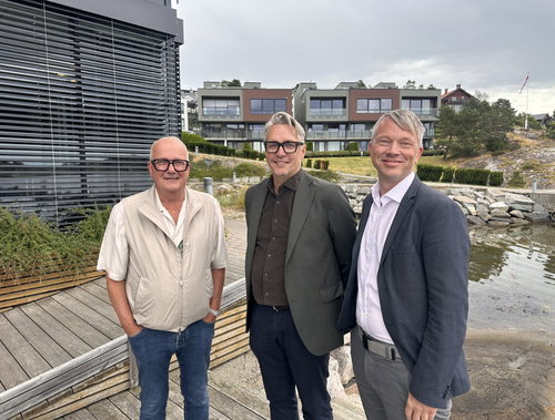 Three men standing outdoors with a cityscape and riverside in the background, smiling at the camera